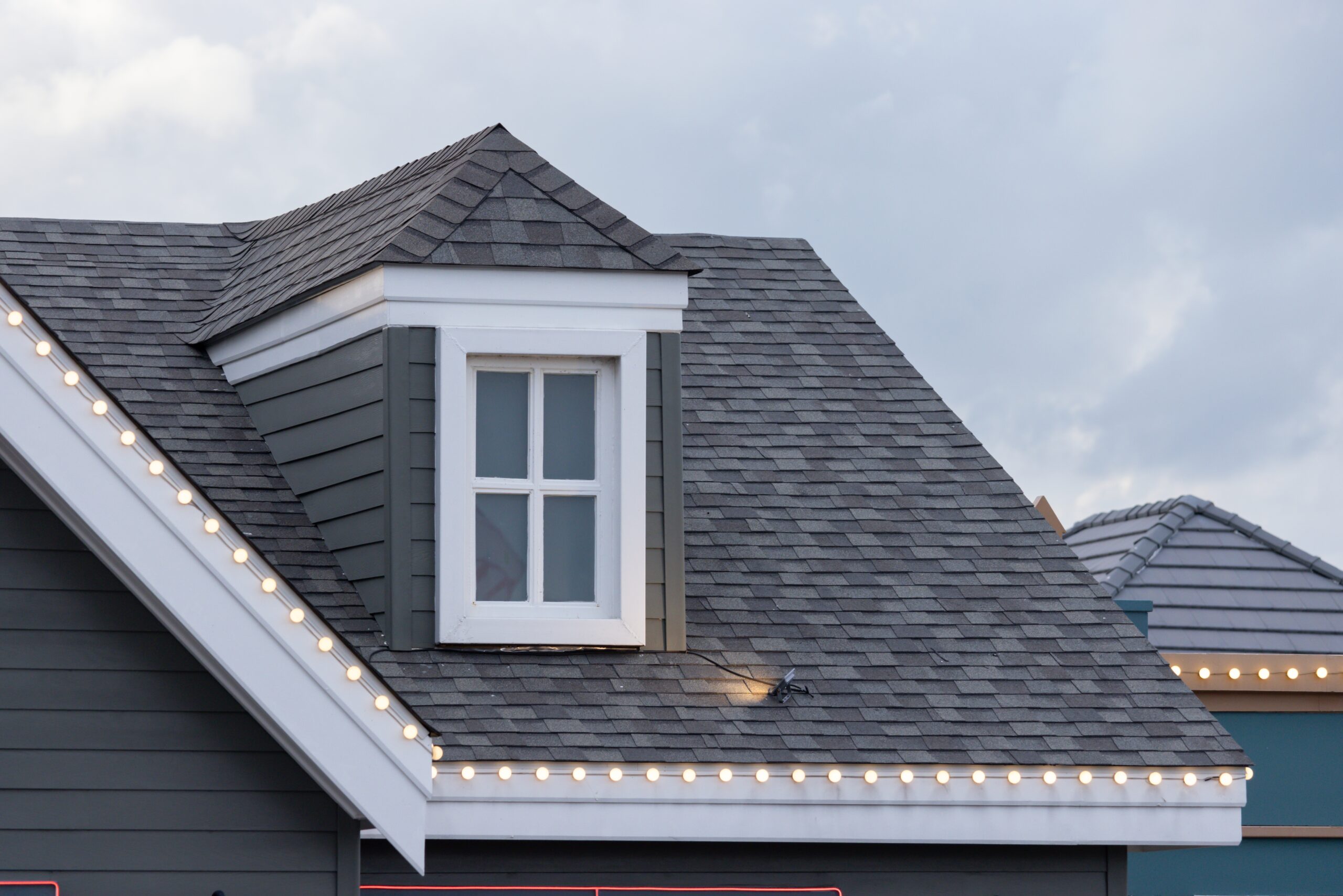 Close-up of a gray shingle roof with a dormer window and decorative string lights along the edge.