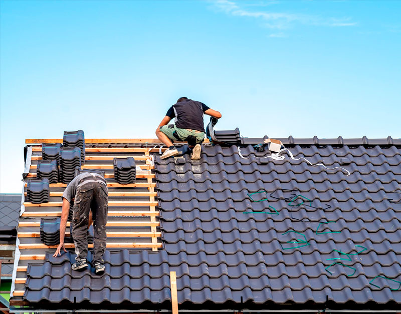 Two workers installing black clay roof tiles on a sloped wooden frame.